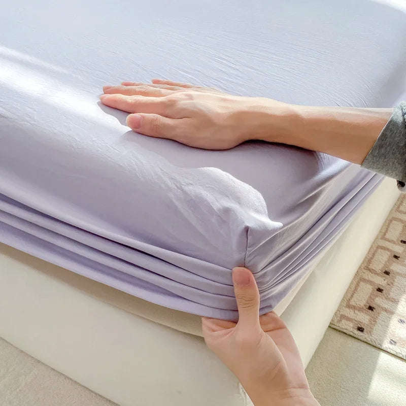A hand is folding a purple fitted bed sheet on a white bed.