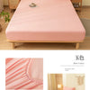 A pink fitted bed sheet set is displayed on a wooden bed frame with pillows and bedside lamps.
