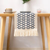 A blue and white geometric patterned linen cotton table runner with tassels displayed on a wooden table.