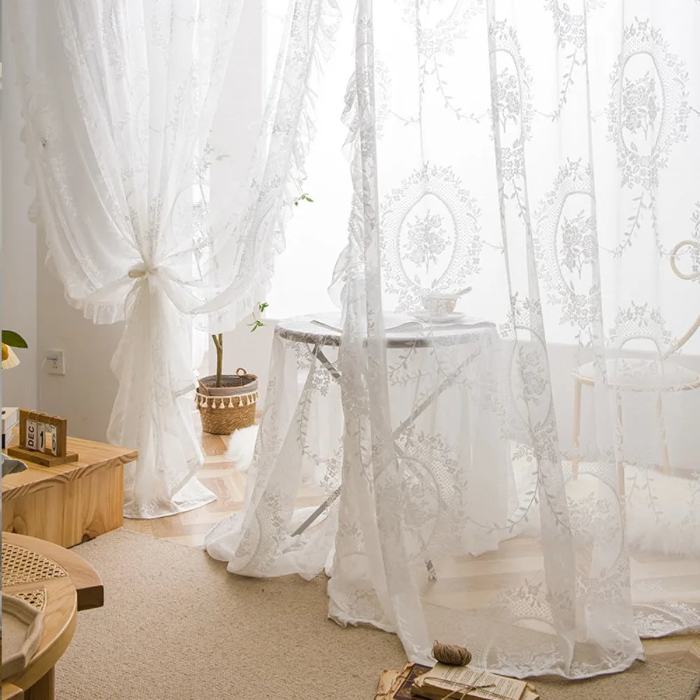 A sheer white floral lace curtain set draped in a window, with a table covered by the fabric.
