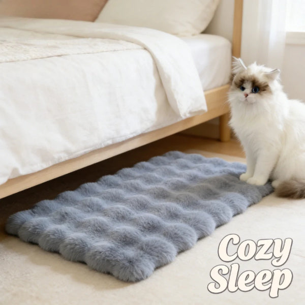 A fluffy gray pet mat is placed on a light floor next to a white cat and a bed.