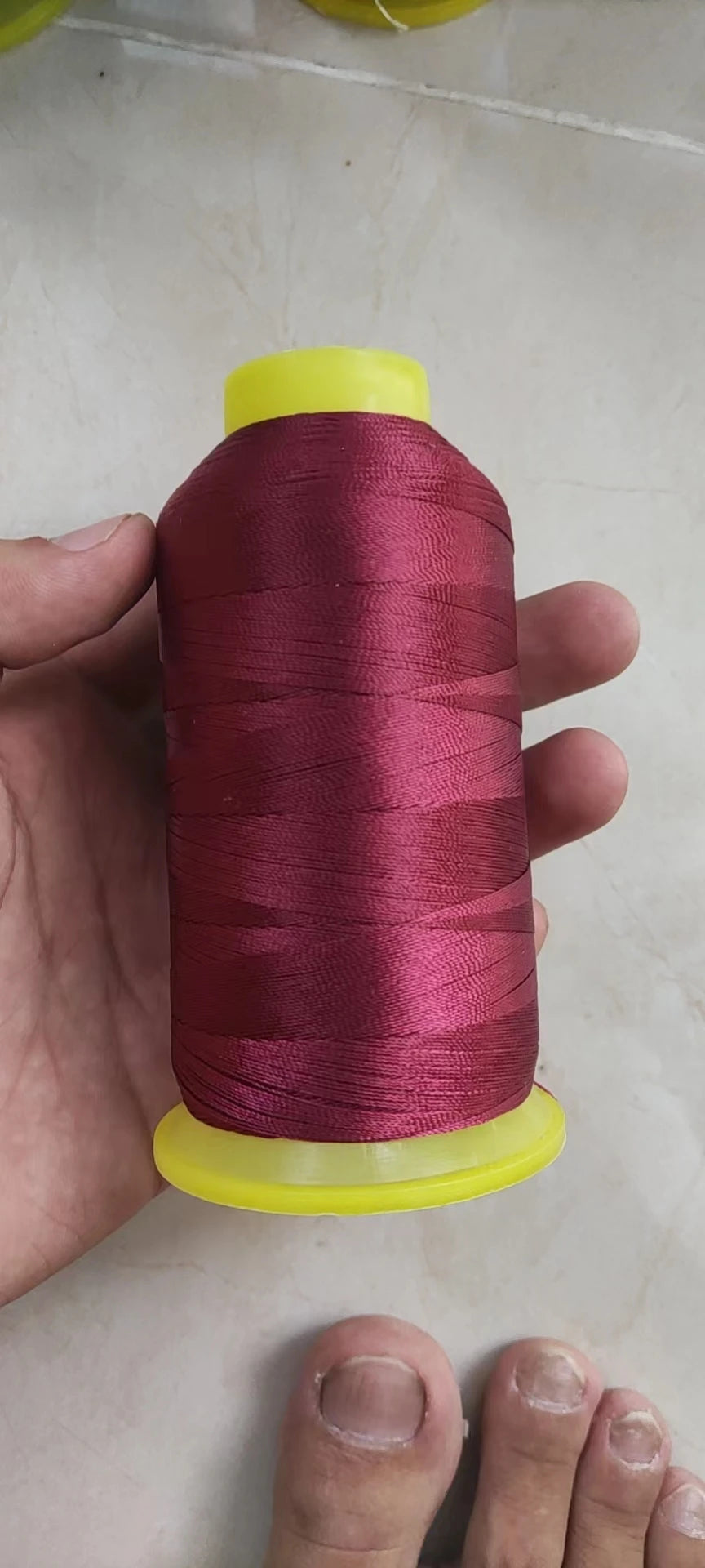 A hand holds a spool of magenta polyester sewing thread with yellow plastic end caps.