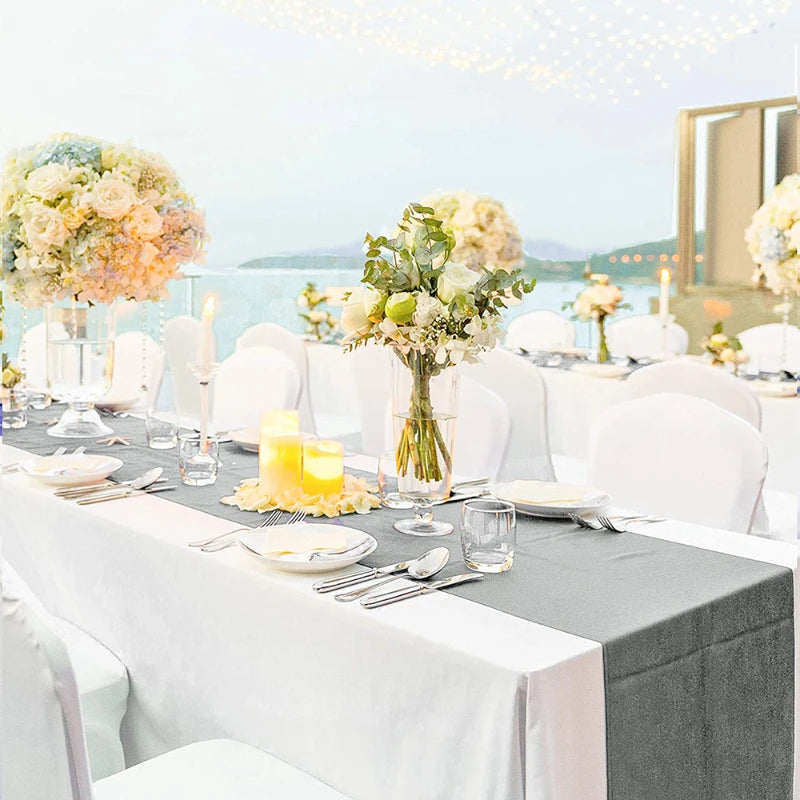 A long table set for an event featuring white chairs, gray linens, floral arrangements, and candles.