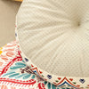 A close-up of a round, light beige cushion with a textured surface and decorative stitching resting on a colorful patterned f