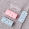 Three non-slip bed sheet grippers in pink, clear, and light blue are displayed on a fluffy gray surface.