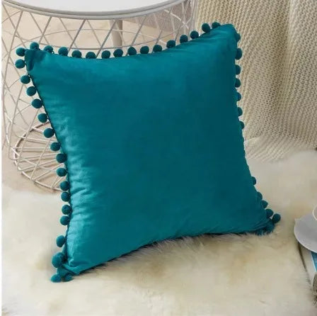 Teal velvet decorative cushion cover with a fringe trim resting on a white mesh table and a fluffy surface.