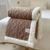 A thick brown fleece blanket with white trim is folded on a white bed, showing a diamond quilted pattern.