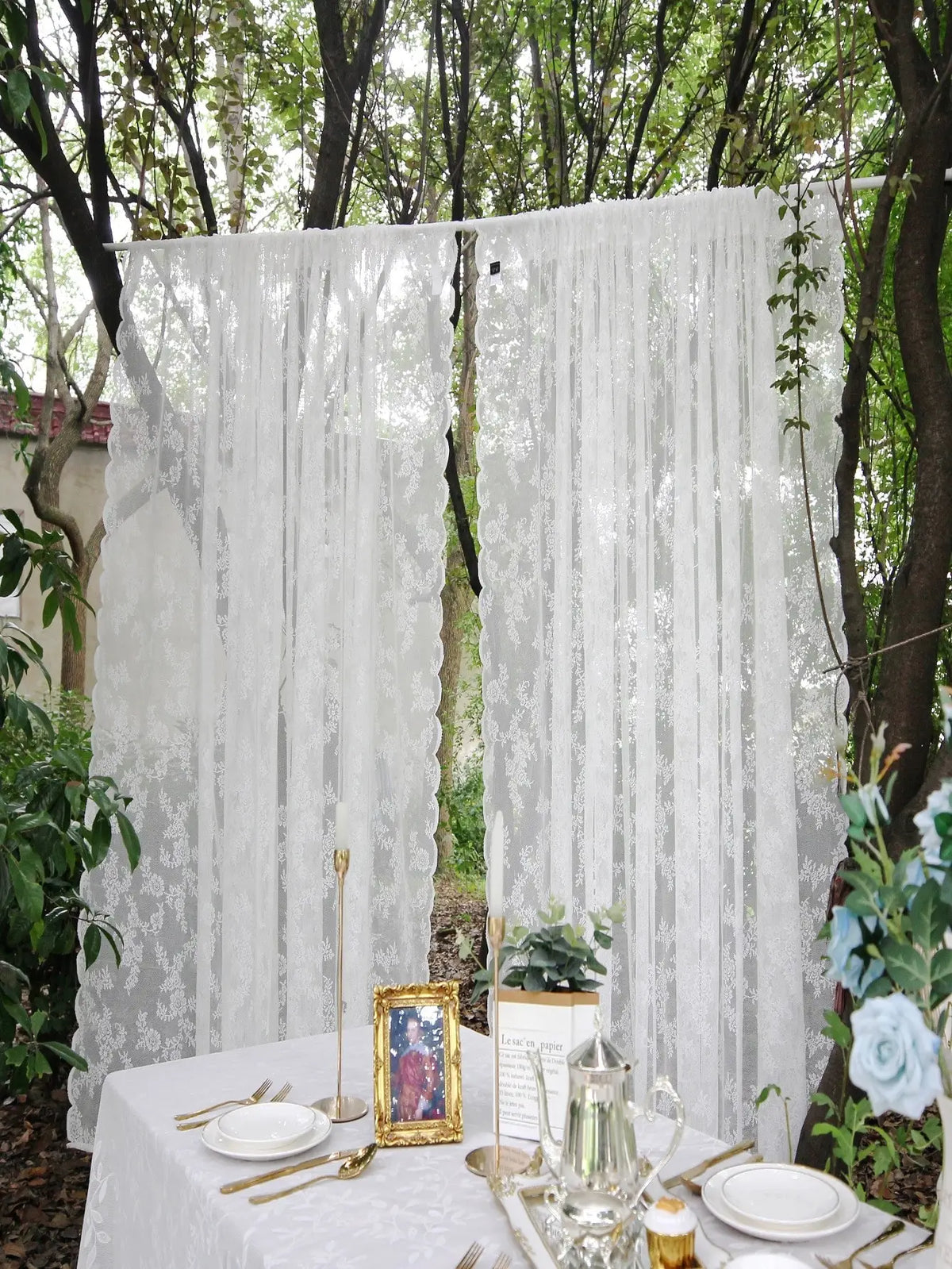A white floral lace sheer curtain panel hanging outdoors in a wooded area.