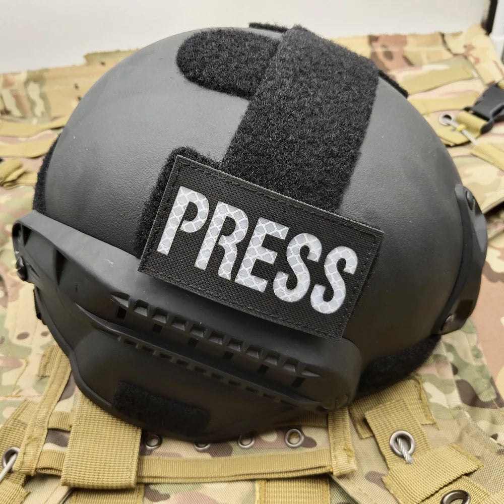 Black tactical helmet with a white PRESS patch on the front, resting on camouflage fabric.