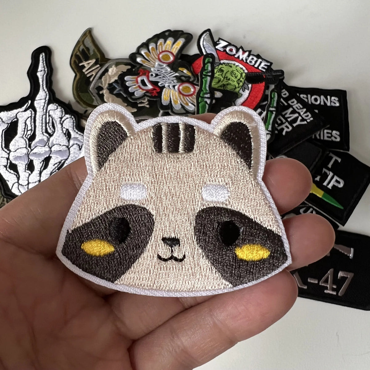 A hand holds a cute panda iron-on patch among various black and gold zombie themed patches.