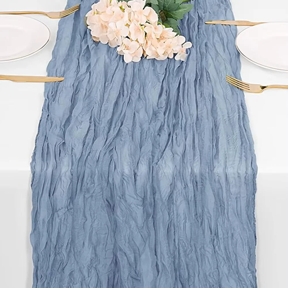 A blue cheesecloth table runner with a textured, wrinkled appearance, featuring pink flowers and gold cutlery.
