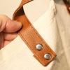 A close-up of a brown leather strap with silver rivets attached to a light canvas fabric.