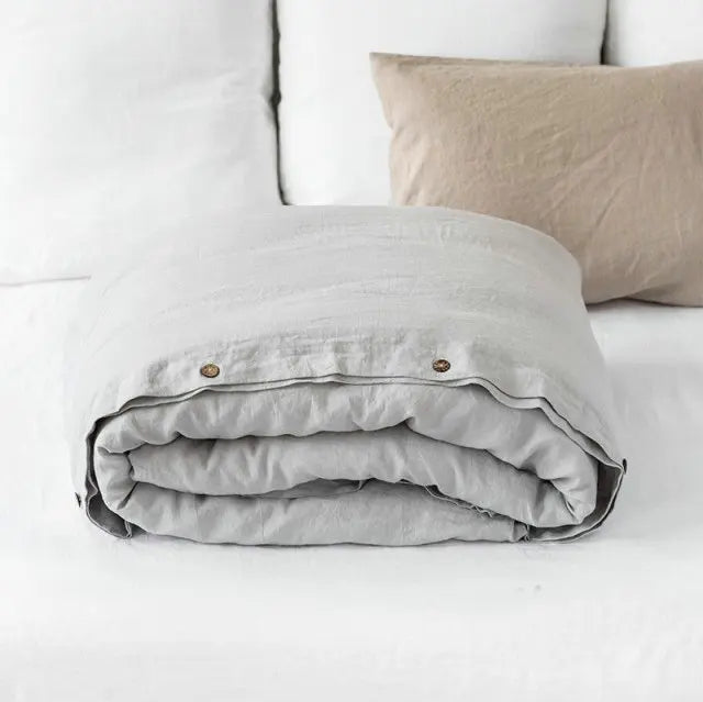 A rolled luxury French linen duvet cover in light gray fabric resting on a white bed with beige pillows.