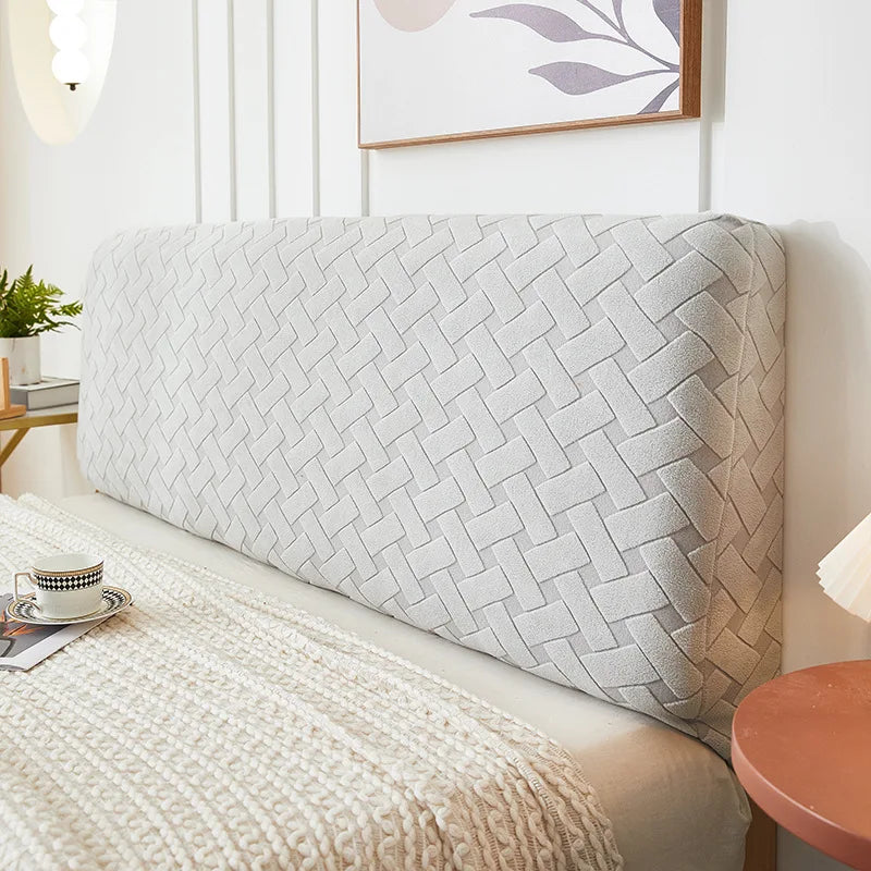 A light gray plush quilted headboard cover is shown on a bed, next to a white wall and framed artwork.