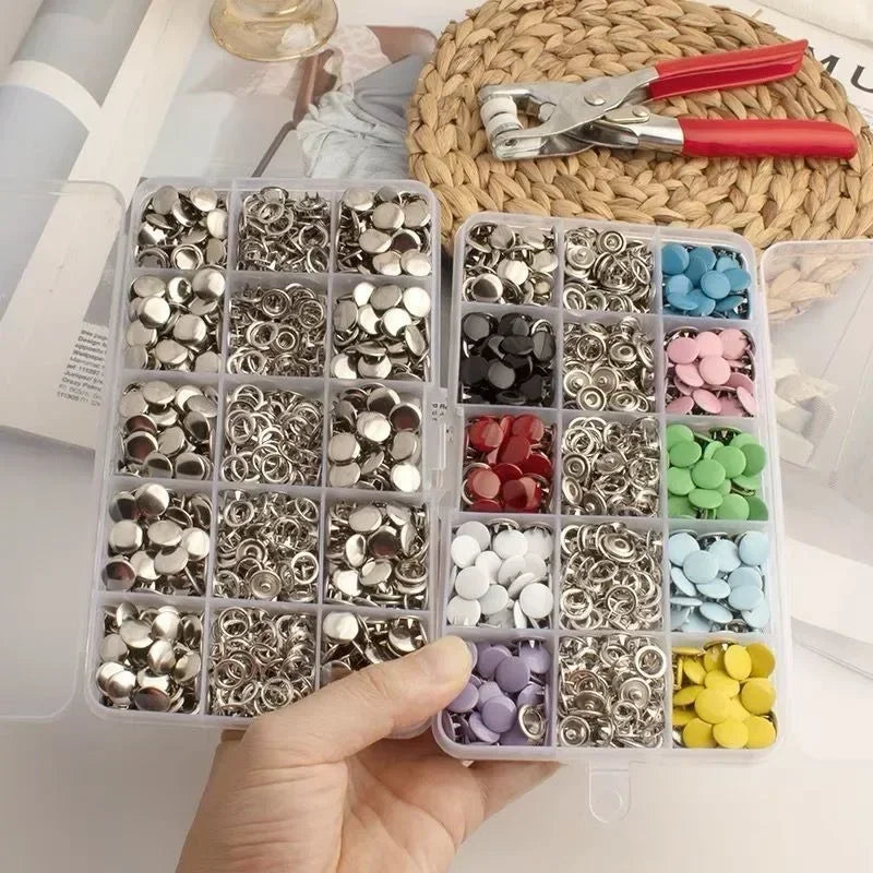 A hand holds an open kit of colorful metal sewing buttons arranged in small compartments on a white surface.