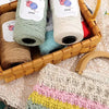 A basket filled with skeins of medium-thick hand-woven cotton yarn in various shades of green, beige, and cream.