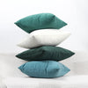 Three velvet cushions in shades of teal, white, and dark green resting on a white textured surface.