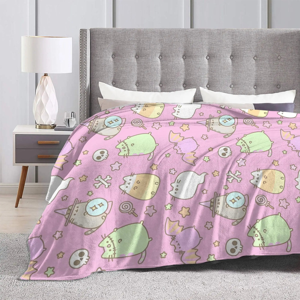 A pink patterned coral fleece cat plush throw blanket draped over a bed with a gray upholstered headboard.