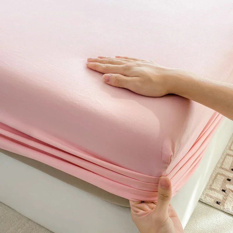 A hand is folding a pink fitted bed sheet made of premium 100% cotton.