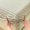 Hands are holding a light beige plaid fitted sheet with a gathered elastic edge.