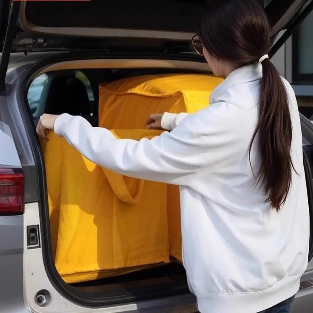 A person is placing a yellow heavy duty foldable storage sack into the trunk of a car.