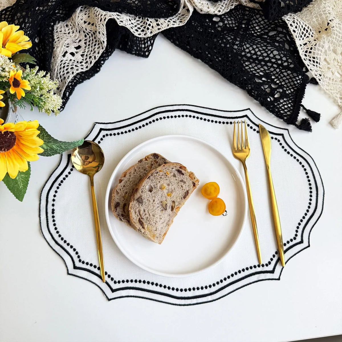 A white plate with sliced bread and gold cutlery is set on a black embroidered linen placemat surrounded by yellow flowers.
