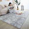 A fluffy gray rug is placed on a light wood floor in a bright room with a sofa and potted plant.
