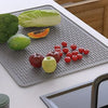 A silicone stove top cover mat is placed on a white surface, holding various fresh vegetables like broccoli, lettuce, and app