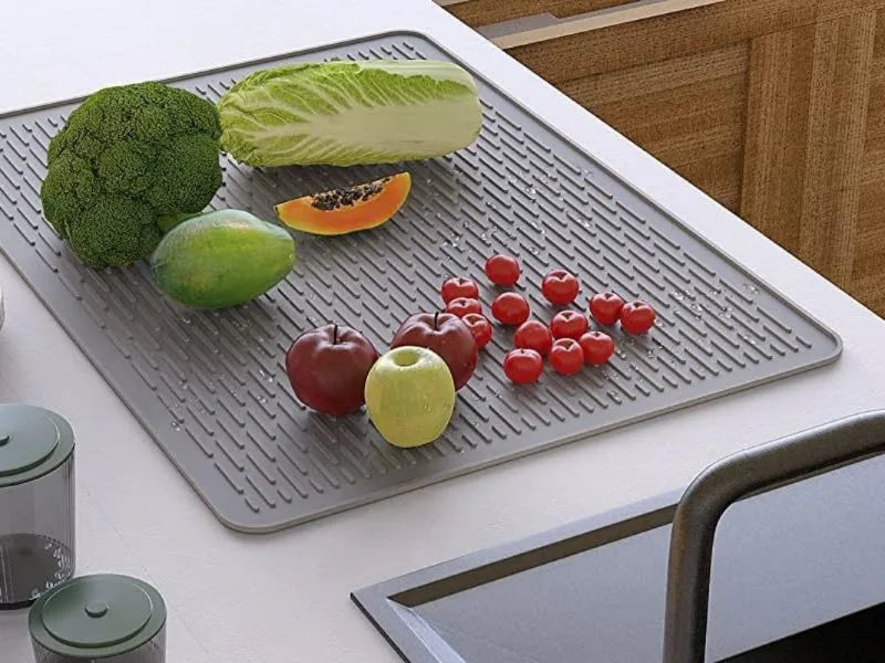 A silicone stove top cover mat is placed on a white surface, holding various fresh vegetables like broccoli, lettuce, and app