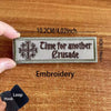 A close-up of a dark gray embroidered patch with the text Time for another Crusade on a wooden surface.