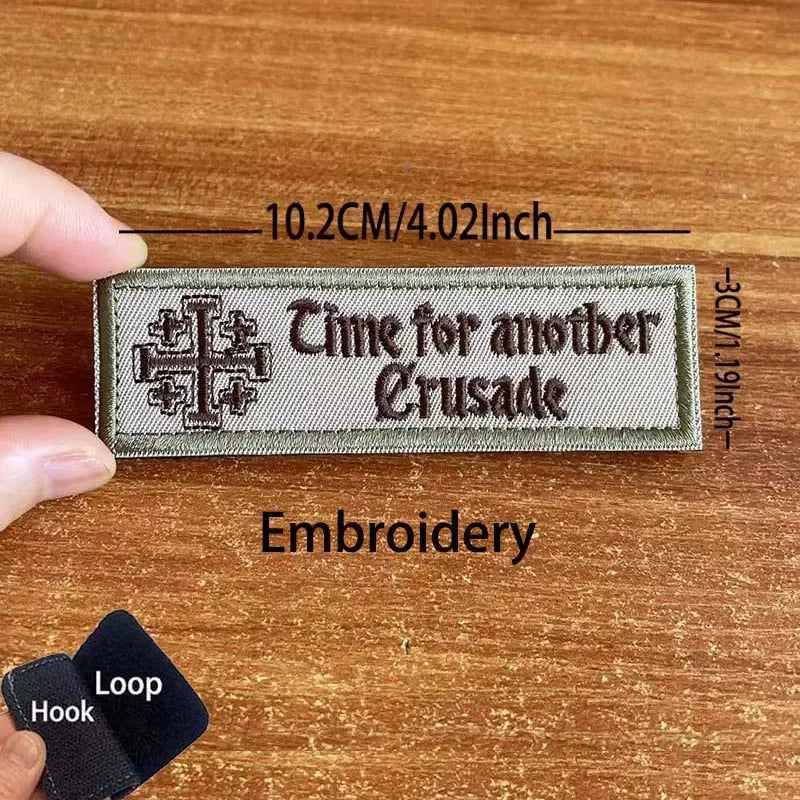 A close-up of a dark gray embroidered patch with the text Time for another Crusade on a wooden surface.