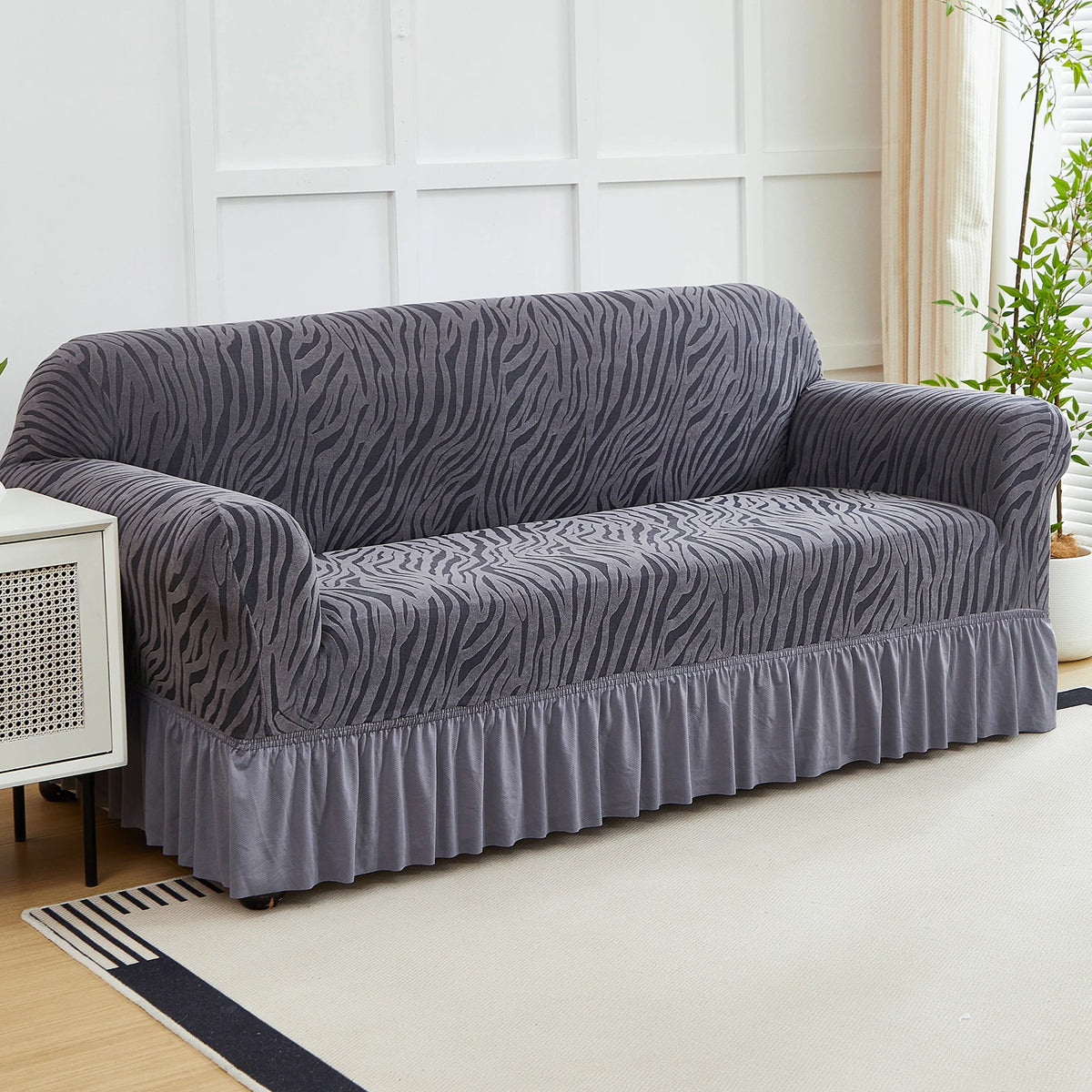 A gray anti-slip jacquard sofa slipcover draped on a light-colored rug in a bright room.