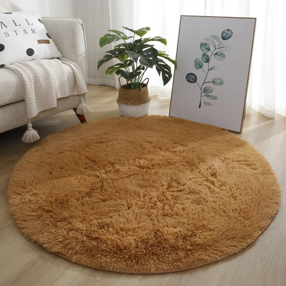 A round, shaggy, golden brown area rug placed on a light wood floor next to a potted plant and framed artwork.