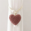 A handmade plush heart tieback in dusty rose color with cream cord and pearl accents, displayed against a white curtain.