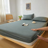 A fitted sheet in a muted blue-gray color is laid out on a wooden bed frame with pillows and a cup of tea nearby.
