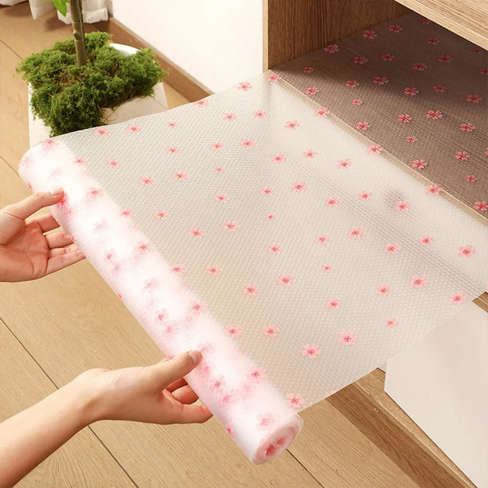 A hand is holding a roll of pink reusable fridge and cabinet liner pads, showing the clear plastic sheet with small floral pa