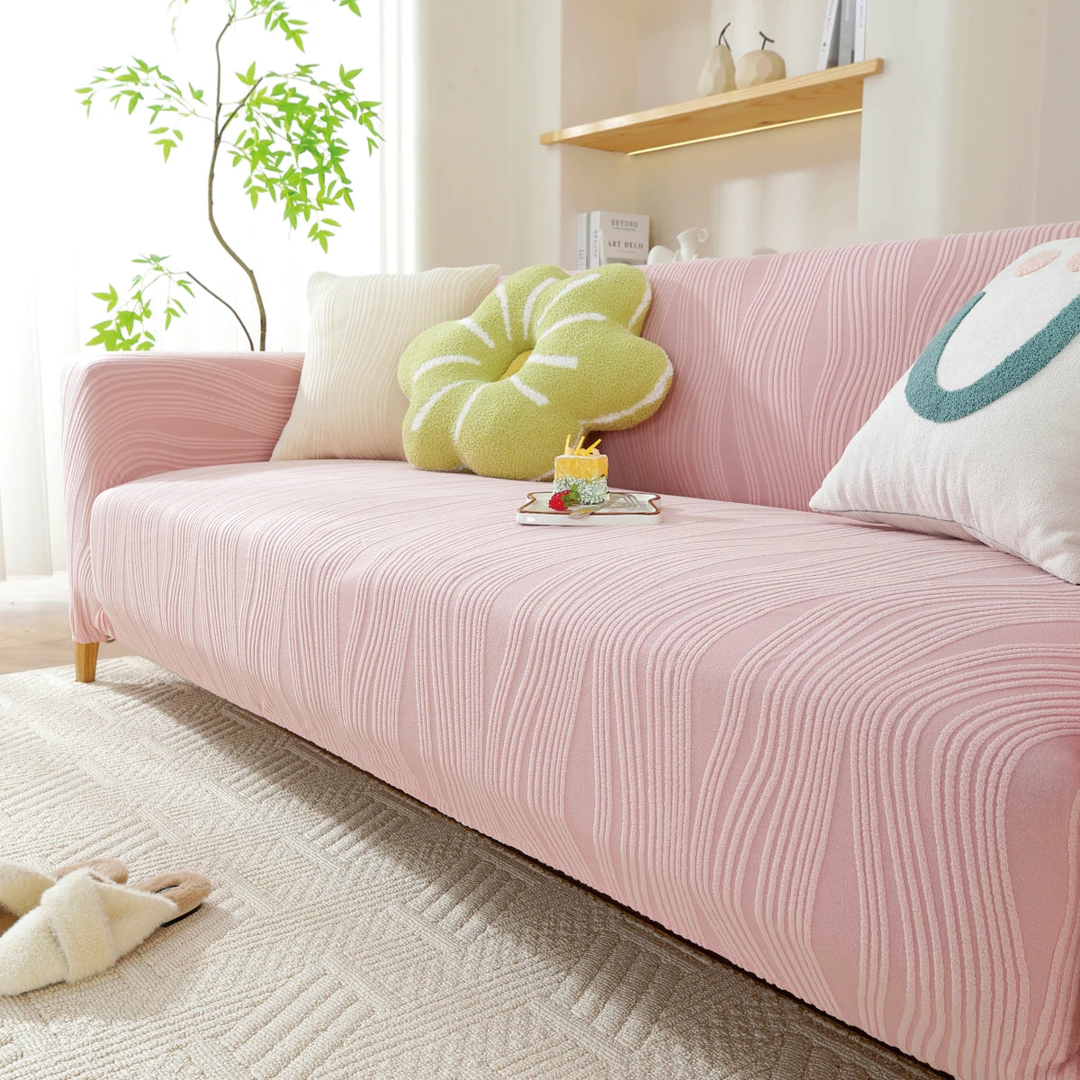 A pink stretchable sofa cover with a vertical line pattern and light green decorative pillows on a beige rug.