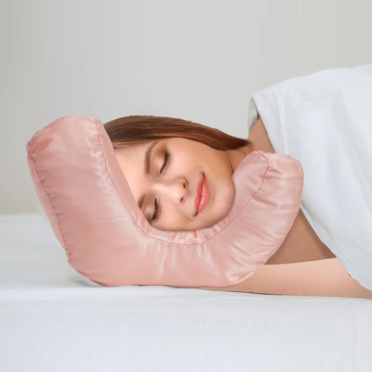 A woman is resting her face on a pink satin anti-wrinkle pillow for beauty and sleep.