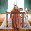 A rustic gauze table runner in dusty rose color draped over a wooden table with natural decor.