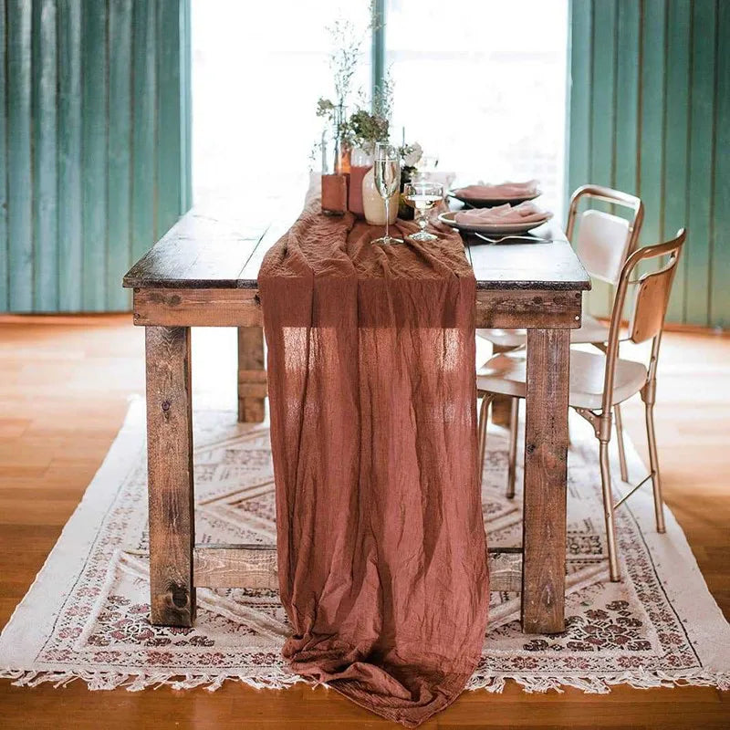 A rustic gauze table runner in dusty rose color draped over a wooden table with natural decor.