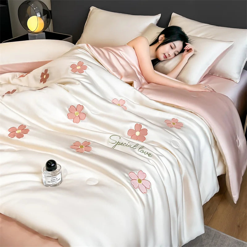 A woman is relaxing under a light pink and cream embroidered quilt in a bedroom setting.