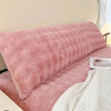 A pink plush long pillowcase resting on a bed, with a white textured blanket nearby.