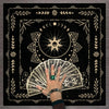 A hand holds a fan of tarot cards within a decorative mat featuring floral and celestial designs.