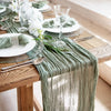 A rustic sage green gauze table runner decorated with white flowers and glassware on a wooden table.