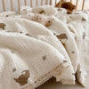 A white muslin blanket with a subtle floral print featuring small animal motifs is folded and displayed next to soft stuffed 
