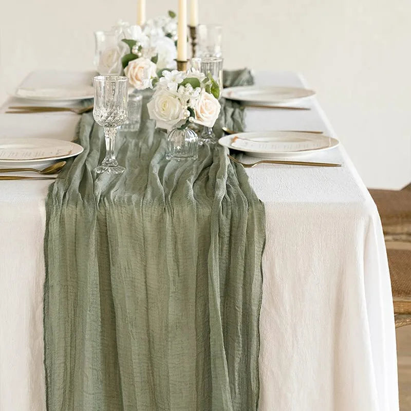 A rustic sage green gauze table runner decorated with white flowers and candles on a white tablecloth.
