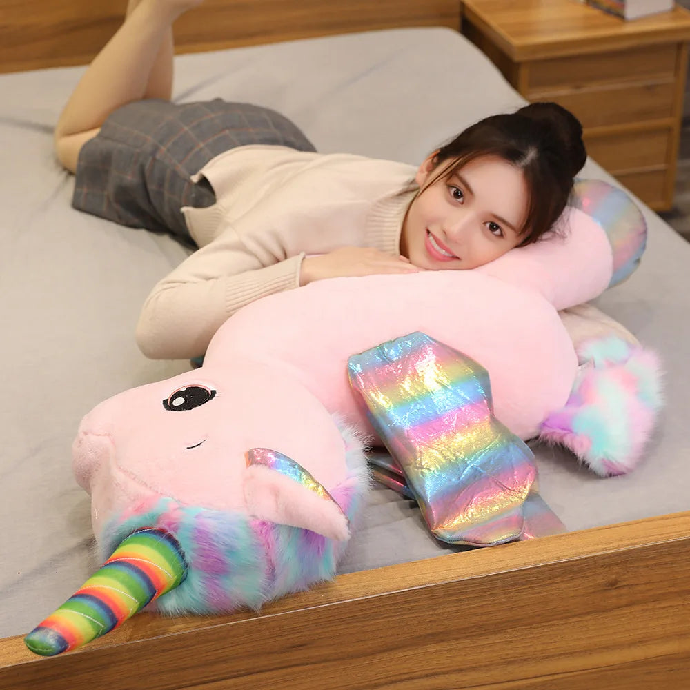 A woman is relaxing on a bed with a pink unicorn plush toy and a rainbow-colored plush toy.