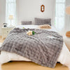 A gray faux rabbit fur throw blanket draped over a white bed with light gray pillows and natural light.