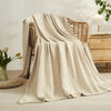 A beige ultra-soft 100% cotton gauze blanket draped over a wicker chair in a bright, minimalist room.
