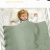 A baby is sleeping peacefully under a soft sage green cotton blanket in a white crib.